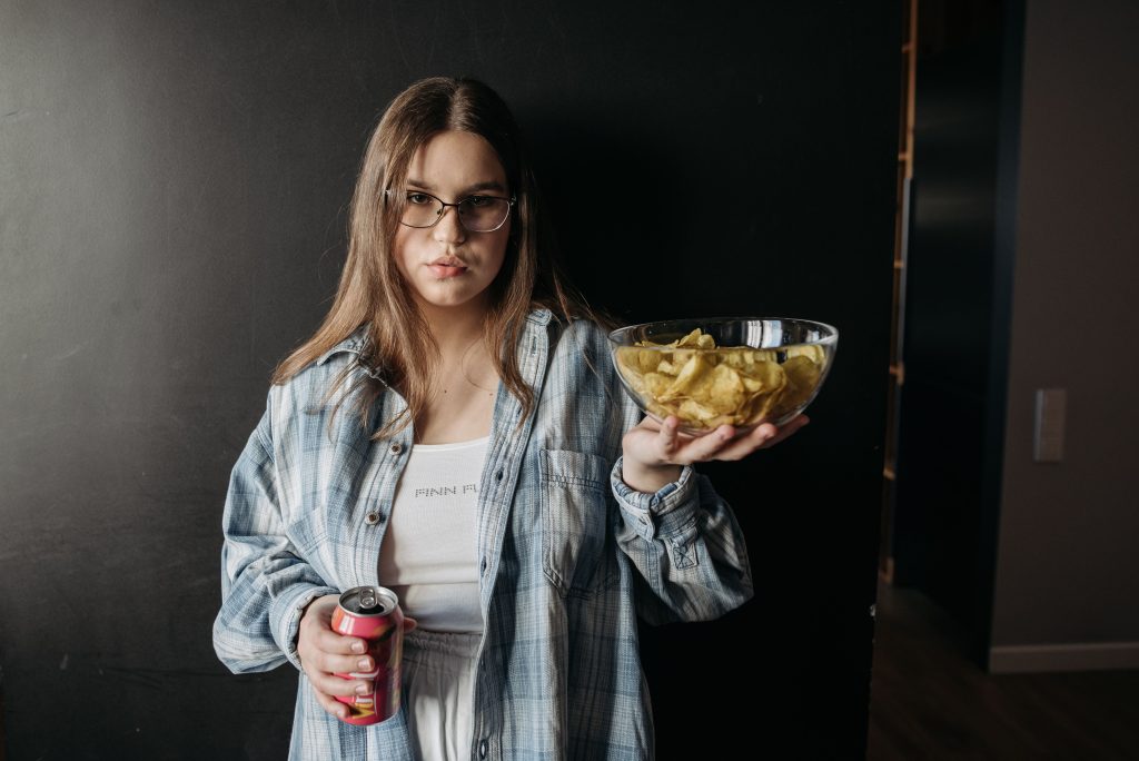 Casual woman holds chips and soda, posing indoors against a dark background.